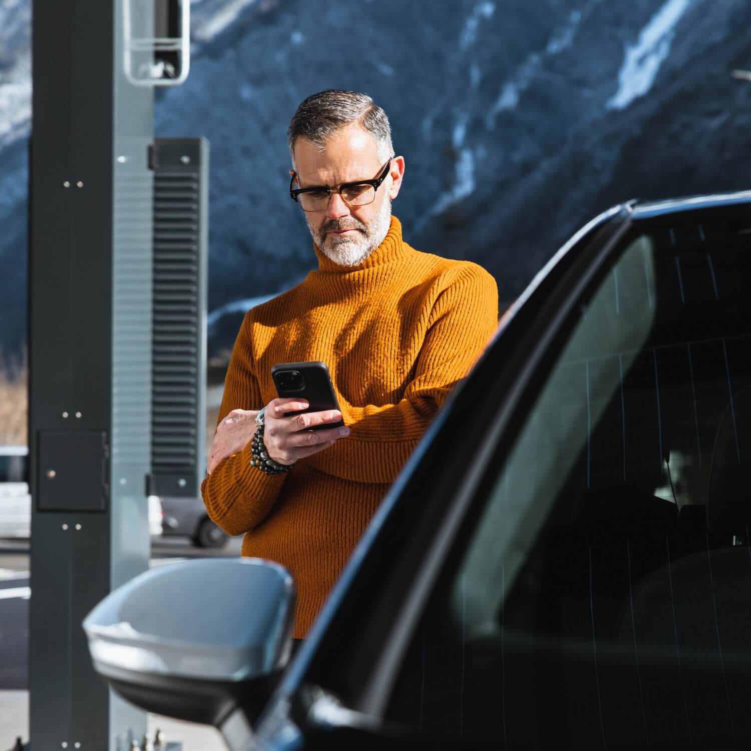 Man im orangefarbenen Pullover am Elektroauto mit Smartphone an Ladestation